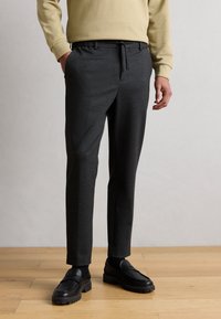 Γκρι παντελόνι με στυλ κωνικό και ρυθμιζόμενη μέση με κορδόνι, κατασκευασμένο από απαλό ύφασμα, σε συνδυασμό με μαύρα μοκασίνια πάνω σε ξύλινο πάτωμα.