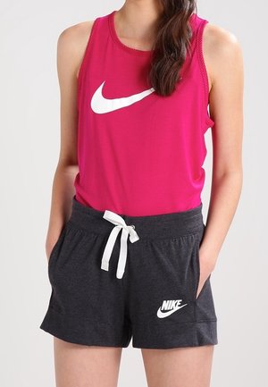 Femme portant un débardeur Nike rose vif et un short Nike gris foncé avec un cordon blanc, debout les mains dans les poches.