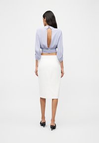 Γυναίκα που φοράει ανοιχτόχρωμο μωβ μπλούζα με ανοιχτή πλάτη και λευκή φούστα μέχρι τα γόνατα, στέκεται με την πλάτη προς τα εμπρός σε λευκό, απλό φόντο.