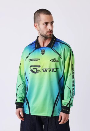 Hombre con cabello corto que lleva una camisa de manga larga con un degradado en verde y azul con los textos "Revolt" y "Change the Game", mirando hacia un lado.