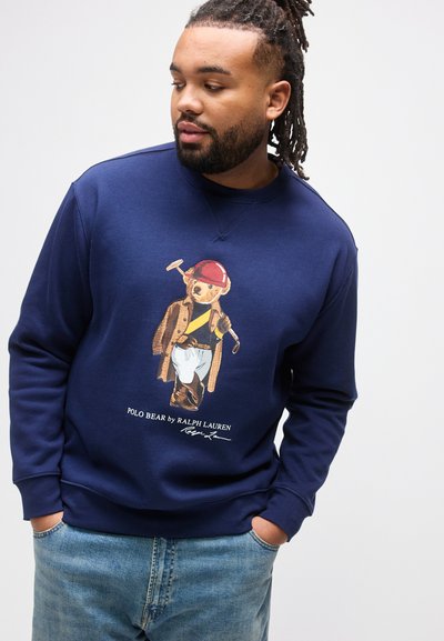 Άνδρας με dreadlocks που φοράει μπλε φούτερ με το σχέδιο ενός αρκούδου Polo ντυμένου με εξοπλισμό πόλο, σε συνδυασμό με ανοιχτόχρωμο μπλε τζιν, με τα χέρια στις τσέπες.