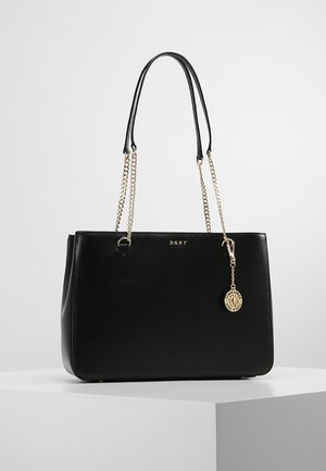 Czarna torba DKNY typu tote z złotymi uchwytami w formie łańcuszków, złotym breloczkiem z logo oraz gładką skórzaną fakturą na białym postumencie na szarym tle.