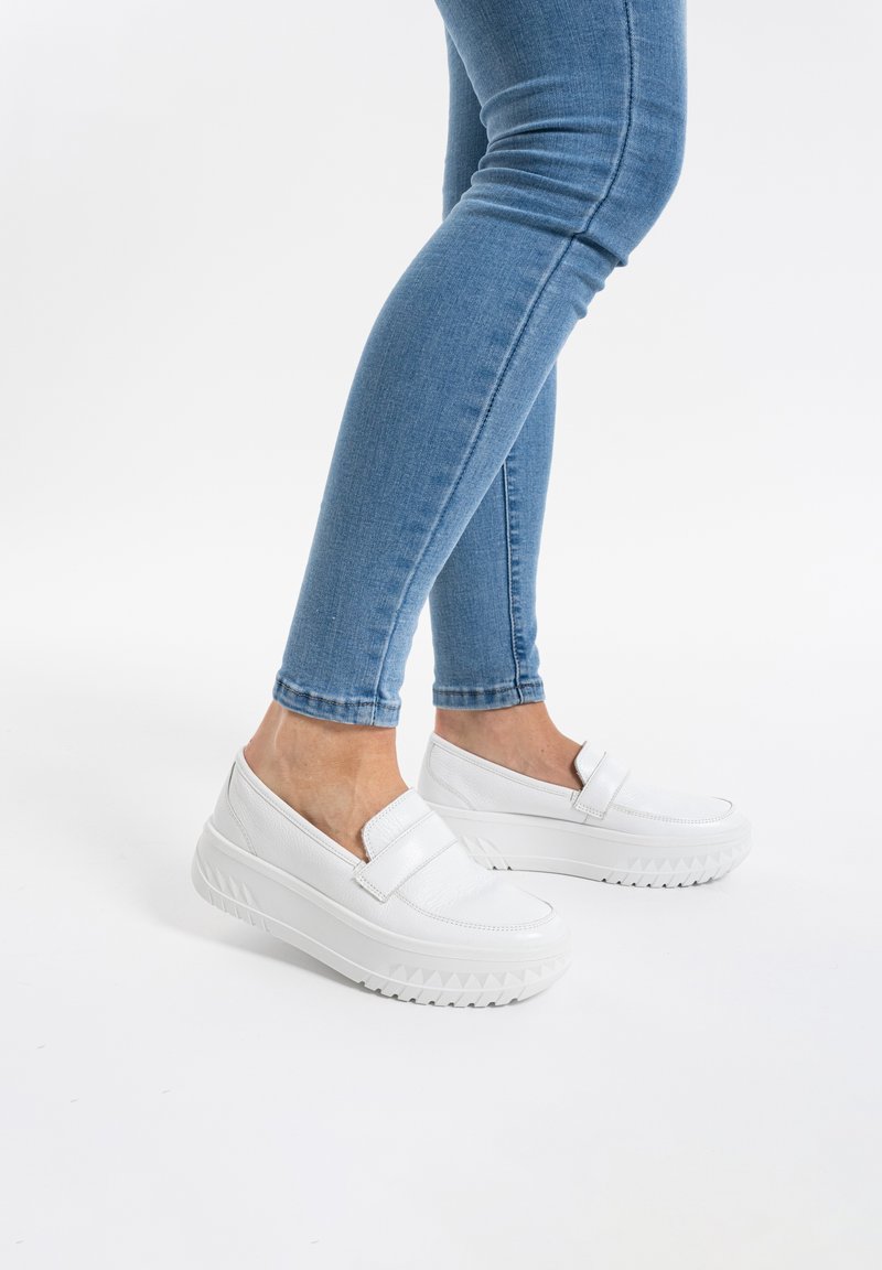 White slip-on shoes with a smooth leather texture, elevated platform, and zigzag pattern on the sole, paired with blue skinny jeans.