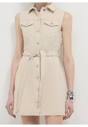 Sleeveless beige button-front dress with collar, tie belt at waist, chest pockets, and large silver bracelet on right wrist.