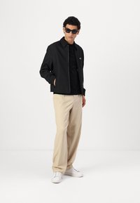 Black lightweight jacket with a straight cut, worn over a black shirt, paired with beige trousers and white sneakers. Bold dark sunglasses.