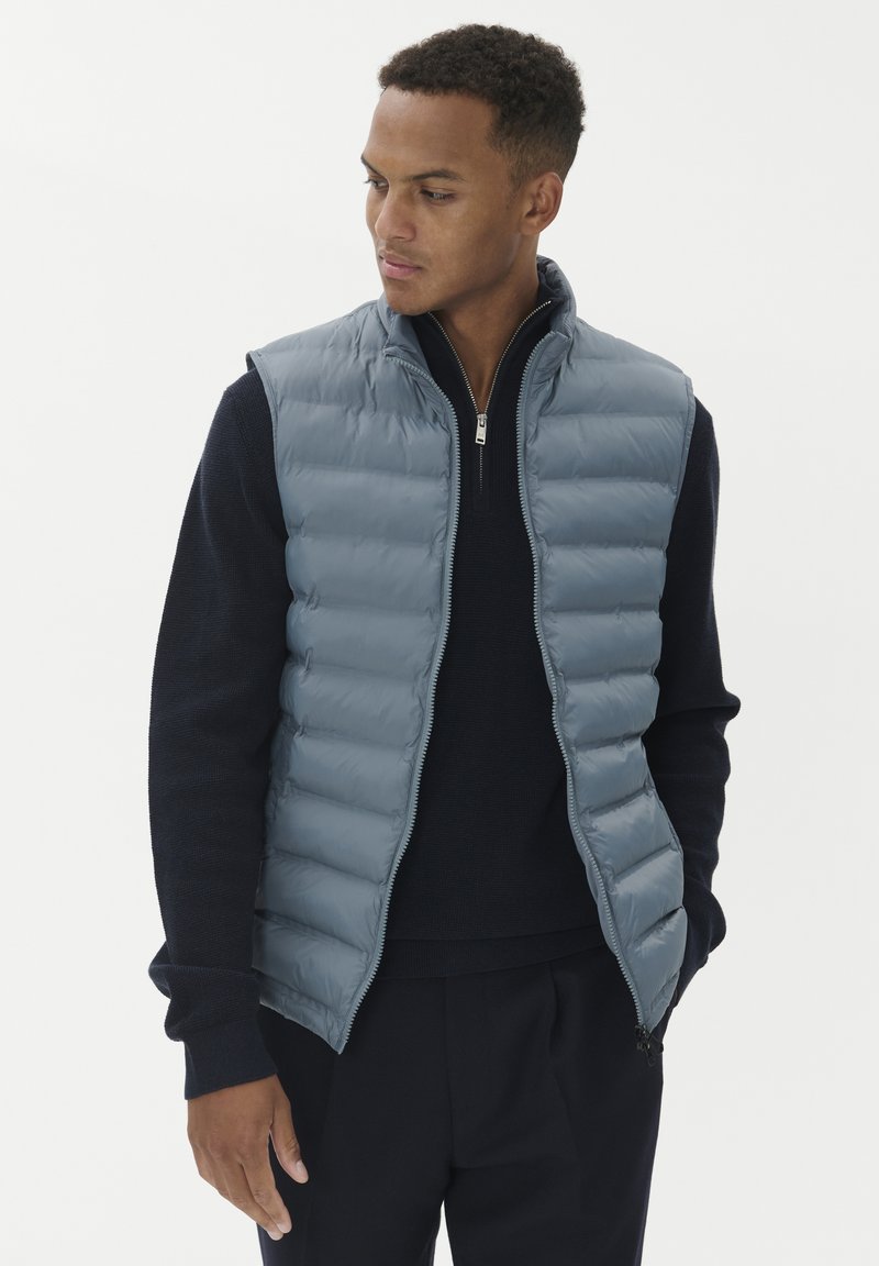 Gai pelēks pūkains vestes ar augstu apkakli, ar rāvējslēdzēja priekšpusi un horizontālu kviltēšanas rakstu, uzlikts virs tumši zila džempera.