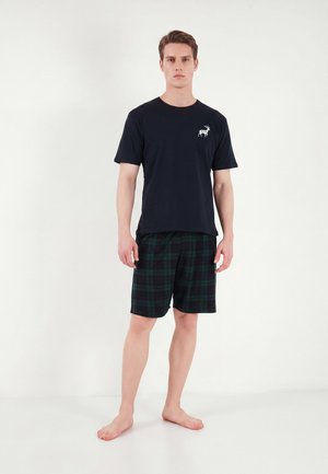 Homme debout pieds nus portant un t-shirt noir avec un logo de cerf blanc et un short à carreaux foncés sur un fond clair uni.