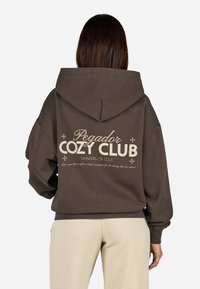 Brauner Kapuzenpulli mit einem lässigen Schnitt, der wei�ßen Text "Pegador Cozy Club" und dekorativen Elementen auf der Rückseite zeigt.