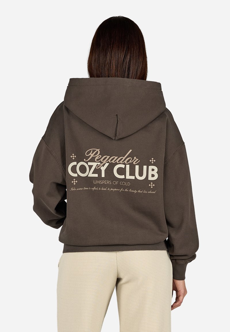 Brauner Kapuzenpulli mit einem lässigen Schnitt, der weißen Text "Pegador Cozy Club" und dekorativen Elementen auf der Rückseite zeigt.