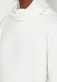 Weißer Rollkragenpullover mit gerippter Struktur, weichem Stoff und umgeklapptem Kragen, entworfen für Wärme und Komfort.