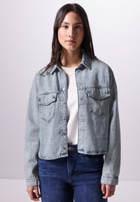 Giacca di denim azzurro chiaro con chiusura a bottoni, due tasche sul petto e design accorciato, abbinata a una maglietta bianca e jeans blu scuro.