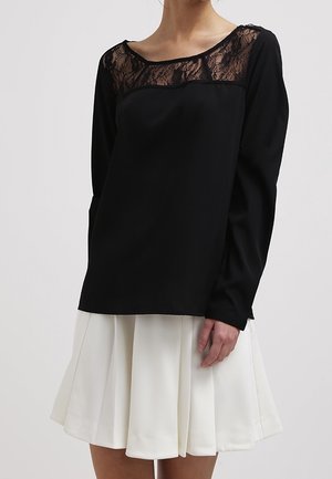 Femme portant un haut noir à manches longues avec un détail en dentelle au niveau de l'encolure et une jupe plissée blanche.