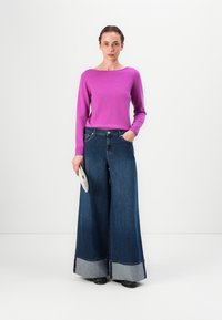 Fioletowy sweter z długim rękawem w połączeniu z szerokimi dżinsami w ciemnym kolorze z jasnoniebieskim mankietem. Modelka trzyma textured torebkę.