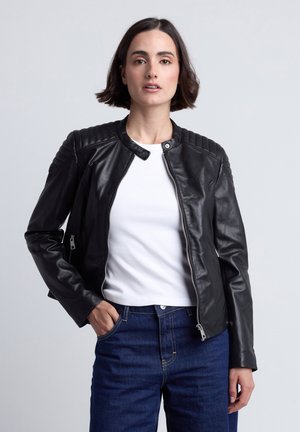 Kvinde med kort mørkt hår iført en sort læderjakke over en hvid skjorte og blå jeans, står med den ene hånd i lommen.