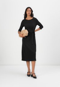 Black, knee-length dress with three-quarter sleeves, belted at the waist; paired with a light brown clutch bag and black strap shoes.