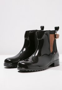 Bottines noires brillantes avec des panneaux latéraux élastiques marron, des tirants, une boucle dorée et des semelles à relief sur une surface blanche.