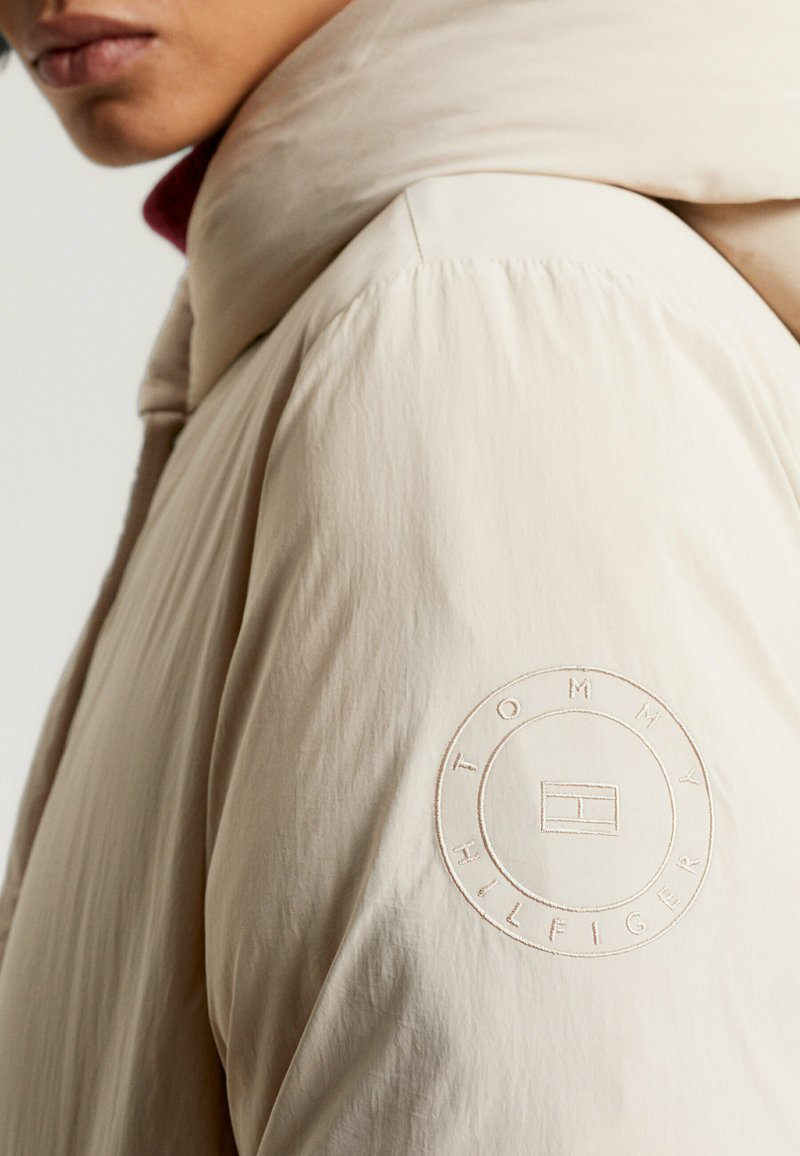 Helle beige Steppjacke mit hohem Kragen, ausgestattet mit einem runden gestickten Logo auf dem linken Ärmel und einem glatten, strukturierten Stoff.