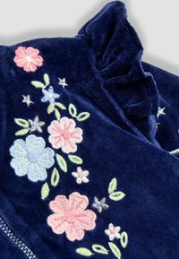 Prenda de terciopelo azul marino con flores bordadas en rosa, azul y verde. Detalle de escote con volantes y cierre de cremallera en la parte frontal.
