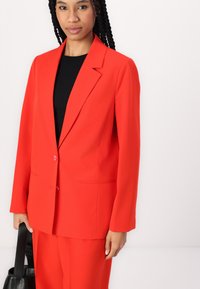 Felrood blazer met een ingesneden revers, enkele knoopsluiting en twee voorzakken. Gedragen over een zwarte top, gecombineerd met bijpassende broek.