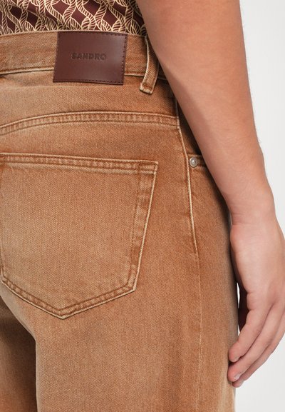 Brune denim jeans med et lædermærke, der står "SANDRO." Funktioner inkluderer en baglomme og synlige sømme på taljen.