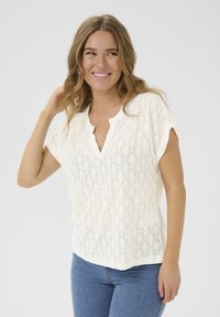 Hvid kortærmet bluse med et struktureret diamantmønster og en delvis knaplukning. Kombineret med tætsiddende blå jeans.