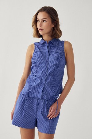 Vrouw draagt mouwloos blauw ruitjesblouse met knopen en gerimpelde zijkanten, gecombineerd met bijpassende korte broek, staand tegen een effen lichte achtergrond.