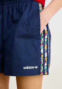 Marineblaue Adidas-Shorts mit floral gemusterten Streifen, elastischem Bund, Seitentasche und Hand teilweise in der Tasche.