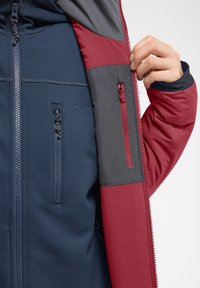 Veste isolante navy et rouge avec un panneau gris. Comprend des zips sur la poitrine et la manche, ainsi qu'un logo de marque visible. Matériau lisse et respirant.