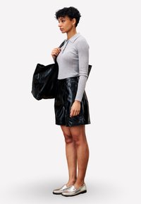 Vrouw met kort, krullend haar, gekleed in een grijs langemouwen poloshirt, zwarte leren rok, zilveren loafers, die een grote zwarte schoudertas draagt en zijwaarts staat.