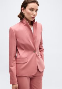 Γυναίκα με κοντά καστανά μαλλιά που φοράει ροζ σακάκι ραμμένο στα μέτρα και ταιριαστό παντελόνι, στέκεται με τα χέρια στις τσέπες μπροστά σε απλό φόντο.
