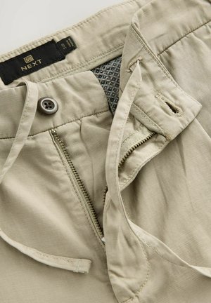 Béžové bavlnené nohavice so zipsovým zapínaním a gombíkovým uzáverom. Obsahujú vnútorný látkový detail s geometrickým vzorom a pútka na opasok.
