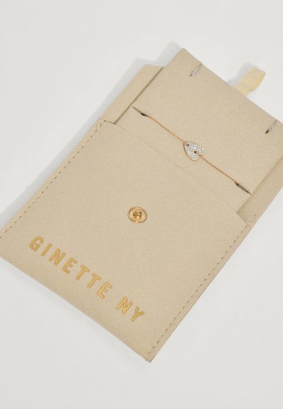 Beige bőrszerű csomagolás arany betűzéssel, amely egy finom arany lánc karkötőt tartalmaz, szív alakú charm-mal, amely gyémántokkal van díszítve.