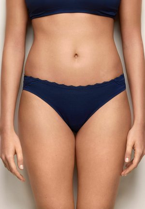 Mujer con bragas azul marino con encaje y sujetador a juego, mostrada desde la cintura hasta la parte superior de los muslos contra un fondo neutro.