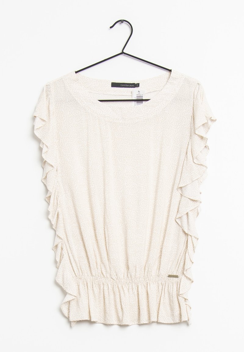 Haut beige clair avec des manches volantées, taille élastique et motif discret. Accroché à un crochet noir sur fond blanc.