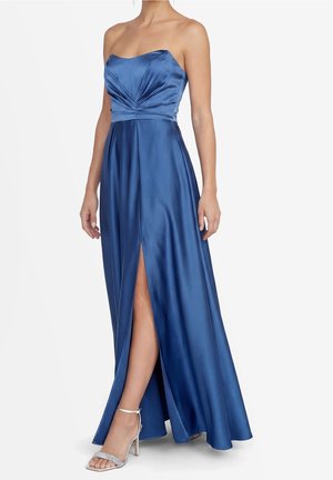 Robe de soirée en satin bleu bustier drapé avec une fente haute à la cuisse, portée avec des sandales argentées à talons hauts.