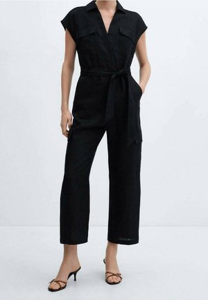 Woman wearing a black sleeveless belted jumpsuit with wide legs and brown heeled sandals, standing against a plain light background.