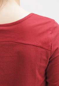 Röd blus med en strukturerad, prickig mönstring. Rundad halsringning och korta ärmar. Tyget verkar mjukt med kontrasterande axeldetalj.