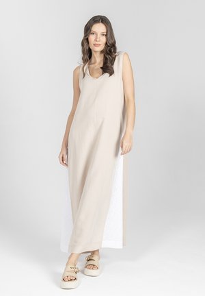 Kobieta o długich brązowych włosach ubrana w beżową sukienkę maxi bez rękawów z białymi panelami po bokach oraz beżowe sandały na platformie, stojąca na szarym tle.