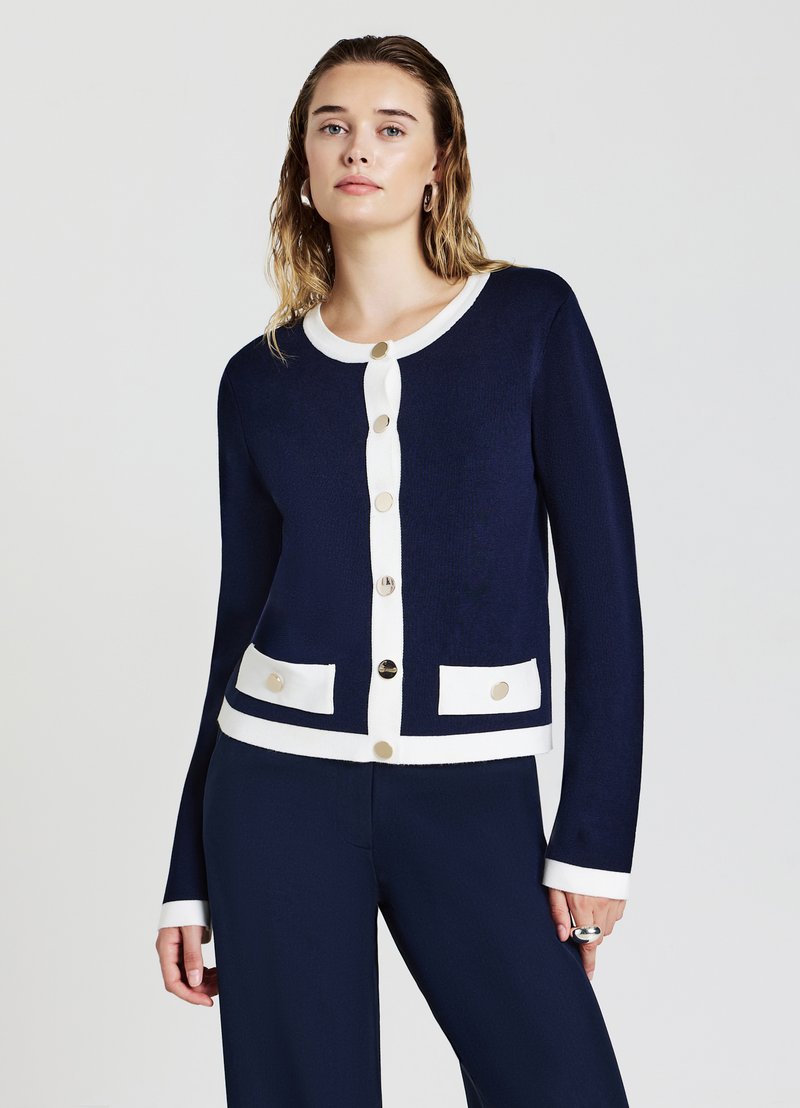 Cardigan di colore blu navy realizzato in materiale a maglia testurizzato, con bordi bianchi, bottoni dorati e due tasche frontali con dettagli a bottone.