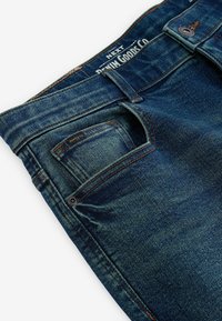 Jeans din denim de culoare albastru închis cu o textură netedă, dotate cu un buzunar frontal, cusături portocalii și un eticheta de marcă pe talie.