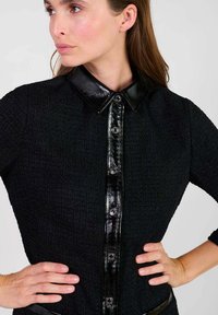 Camisa negra texturizada con un cuello negro brillante y botones frontales, con un diseño ajustado y mangas de tres cuartos.