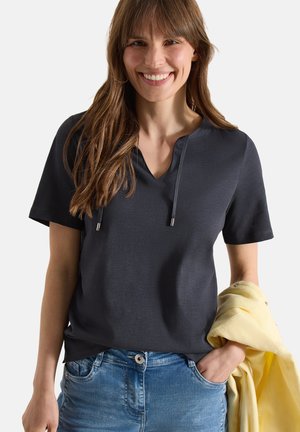 Femme souriante aux cheveux bruns portant un haut gris foncé à manches courtes et un jean, tenant une veste jaune pliée dans sa main gauche.