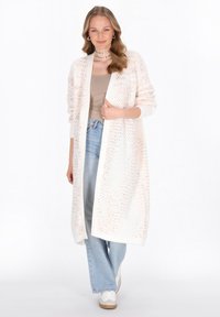 Cardigan lung tricotat, de culoare albă cu stropi pastel, design cu deschidere frontală, manșete cu nervuri și o croială lejeră, purtat peste un top bej și blugi deschiși.