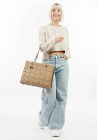Tans gekwantelde neplederen totebag met een rechthoekige vorm, voorzien van zilverkleurige details en een hartvormige charme, gedragen met een casual outfit.
