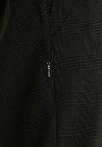 Schwarzer, texturierter Stoff mit einem dezenten Karomuster, an der Seite ist ein kleines Etikett mit der Aufschrift "CHASIN'" angebracht.