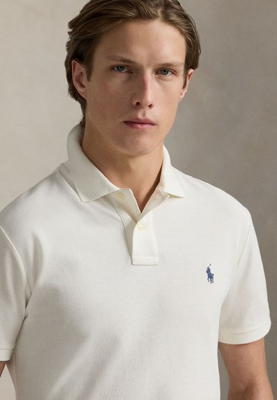 Ung mann med lysbrunt hår iført en hvit polo med liten mørkeblå brodert logo på brystet.