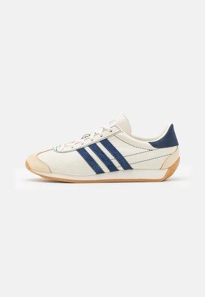 Sneaker in pelle bianca con strisce in camoscio blu, dettagli beige sulla punta e sul tallone, e suola in gomma, mostrato di lato su sfondo bianco.