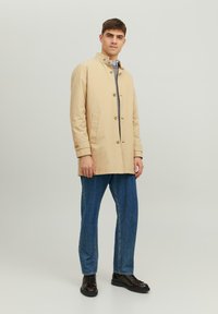Beige katoenen jas met opstaande kraag, knopen aan de voorkant en zijzakken, gecombineerd met blauwe denim jeans en zwarte veterschoenen.