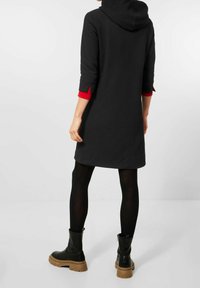 Robe à capuche noire avec manches trois-quarts et poignets intérieurs rouges, réalisée en matière texturée. Associée à des bottines noires et des collants.