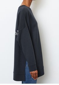 Sudadera de gran tamaño en color gris oscuro con mangas largas, puños acanalados y aberturas laterales. Presenta un detalle de texto en blanco contrastante en el frente.